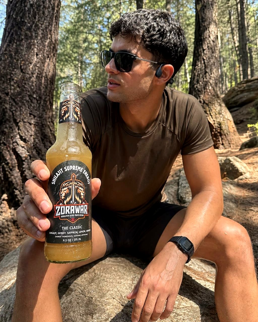A man resting on a rock in a pine forest, holding a bottle of Zorawar